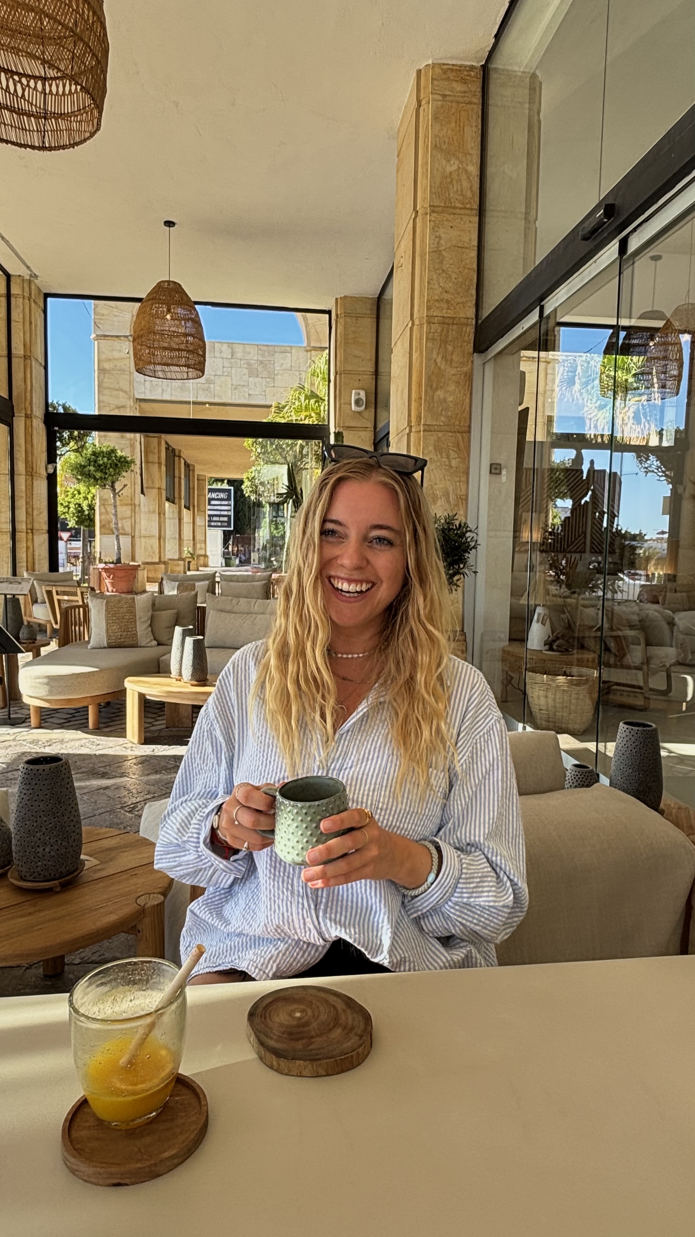 Lächelnde Frau mit langen blonden Haaren, die eine grüne strukturierte Tasse hält, an einem Tisch mit einem Glas Orangensaft in einem sonnigen Außenbereich.